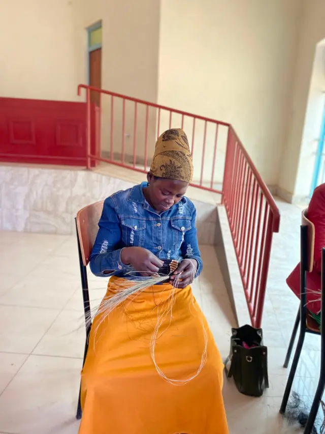 Umugore ukiri muto ufite akantu arimo araboha, yambaye ishati ya 'jeans' akwembetse n'igitambaro cy'umuhondo, yicaye ku ntebe yegamirwa, n'agasakoshi kiwe k'umukara kari hasi ku ibaraza ry'amakaro, hari na griyaje. Ifoto yafotowe muri Nzeri (9) mu mwaka wa 2025. 