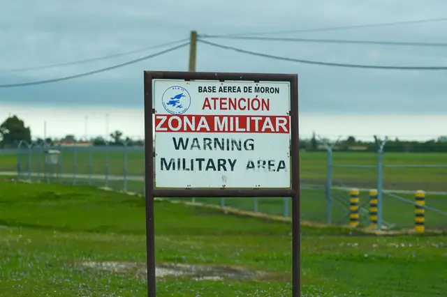 Cartel donde se lee "Base Aérea de Morón. Atención, zona militar. Warning Military Area". Al fondo se ve una llanura verde cercada con vallas. 