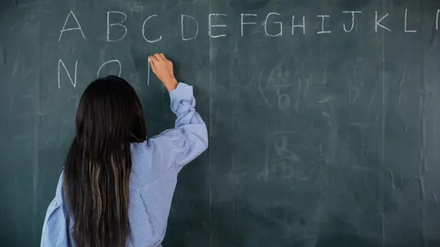 Uma professora escreve com giz na lousa as letras do alfabeto em letras de forma