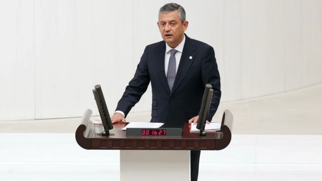 Özgür Özel elini kürsüye koymuş, mecliste konuşma yapıyor. Siyah bir takım giymiş ve karede bir tek o var.