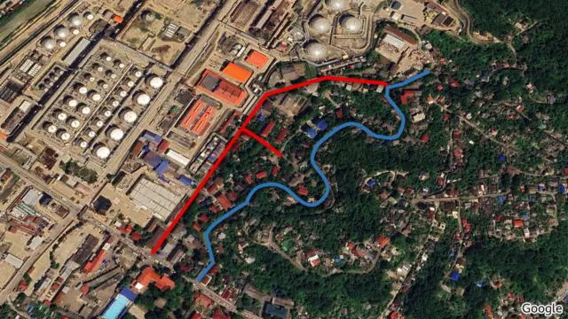 Супутниковий знімок промислової території, зроблений з великої висоти. У лівій частині кадру розташований великий індустріальний комплекс із численними круглими резервуарами та трубопроводами. Праворуч від промислової зони видно житловий сектор з зеленню, садами та деревами. На зображенні додатково нанесено кольорові лінії, що тягнуться вздовж краю промислової зони й частково заходить у житлову забудову.