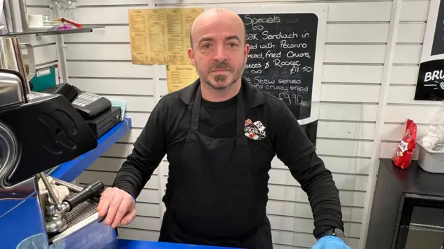 Omar Musu, dueño del café-bar Brushchetta en Filey, y preocupado por la posible repercusión de la prima del impuesto municipal en el empleo, se encuentra detrás del mostrador de su negocio. Su mano derecha está sobre la máquina de café. Detrás de él hay un cartel con los "especiales". Lleva una camiseta negra de manga larga y un delantal negro.