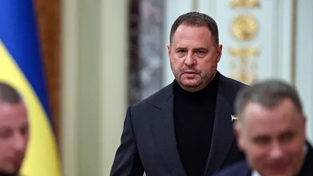 Єрмак в темному діловому піджаку й чорному високому светрі. На лацкані — невеликий значок у вигляді прапорця. Людина йде або стоїть у приміщенні з урочистим інтер’єром: світлі стіни, декоративні елементи з позолотою. На задньому плані частково видно інших людей, розмитих, і синьо‑жовтий прапор ліворуч. 