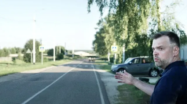 Мужчина в тёмно-синем поло стоит у обочины дороги в деревне в ясный день. Он показывает правой рукой в сторону пешеходного перехода, на заднем плане берёзы, дорожные знаки, столб и серый джип.