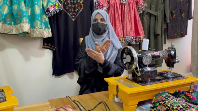 لیدا نومې نجلۍ پيکه بانجاني رنګه ټېکری پر سر، ماسک پر مخ او تور کالي یې اغوستي او د هغه مېز شا ته ناسته برېښي چې د جامو ګنډلو ماشين او ځونډي ښکاري. شا ته یې ډول ډول د ښځو کالي په دېوال کې راځوړند دي. 