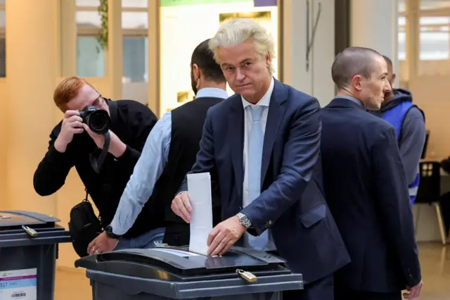 Hollandalı siyasetçi Geert Wilders lacivert takım elbisesiyle oy sandığına oy pusulasını atarken kameraya poz veriyor, bir erkek fotoğrafını çekiyor ve arka planda başka erkekler var