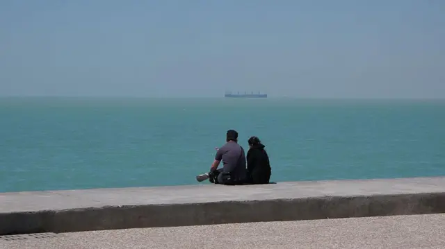 یک زوج جوان هنگام عصر کنار دریا در بوشهر نشسته‌اند، جایی که از دور یک نفتکش در خلیج ‌فارس دیده می‌شود ۱۸ آوریل ۲۰۲۵