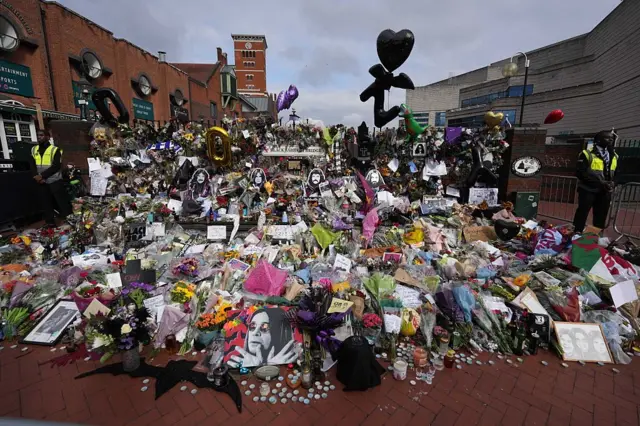 Очень много цветов, черных шаров, портретов Оззи и свечей. В кадре видны два полицейских, стоящим по бокам от фанатского мемориала.