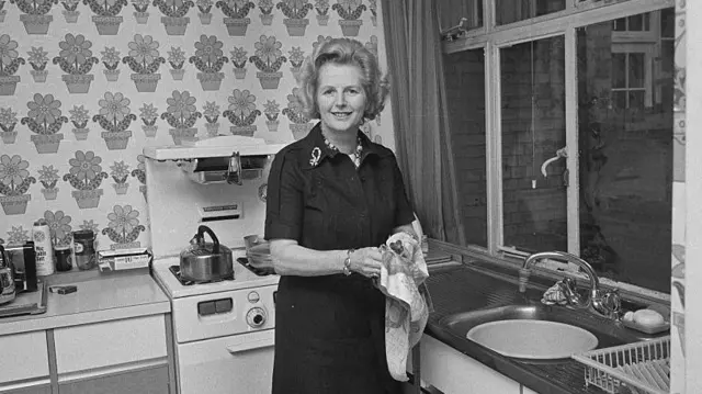 Маргарет Тэтчер стоит возле кухонной раковины с чашкой и полотенцем в руках. На заднем плане плита и чайник