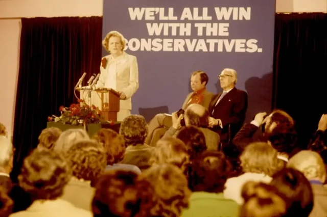 مارگارت تاچر، نامزد نخست وزیری بریتانیا در سال ۱۹۷۹، با شعاری در پشت سر که می‌گوید: «با محافظه‌کاران ما همه برنده‌ایم»