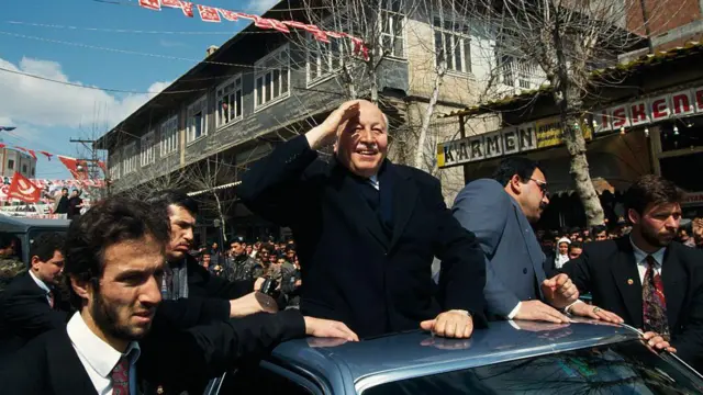 Erbakan bir aracın üzerinden halkı selamlıyor. Etrafta takım elbiseli erkekler ve bayraklar.