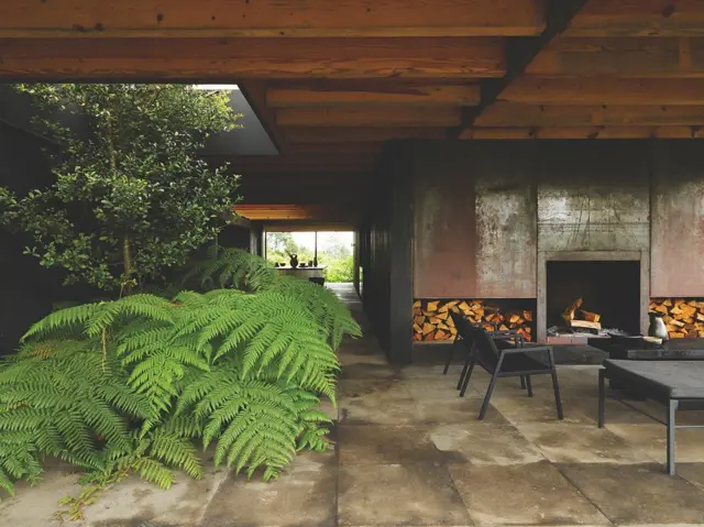 Espacio interior con vigas de madera, plantas frondosas y chimenea moderna integrada en muros de hormigón.