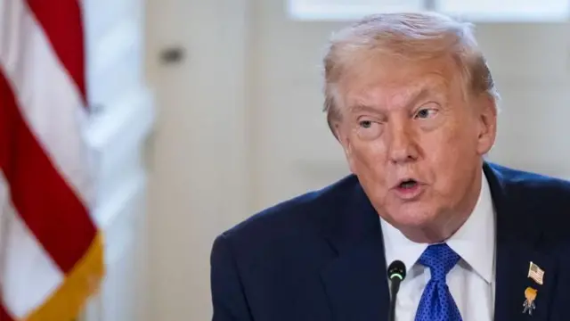 星条旗と白いドアを背に、紺色のスーツ姿のトランプ氏。上着の襟に星条旗のバッジと、トランプ氏をキャラクター化したように見えるバッジをつけている