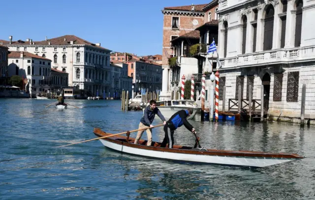 Barco navega21 cassinoVeneza, com dois homens a bordo, um deles com um remo, e casas ao fundo