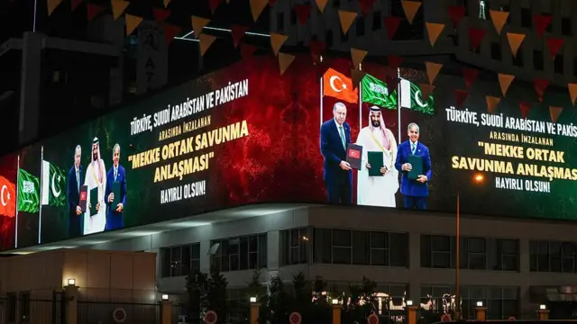 Gece çekilmiş bir fotoğraf. Bina üzerindeki dijital ekranda Erdoğan, Bin Salman ve Şerif ellerinde anlaşma dosyalarını tutuyor. Arkalarında ülkelerinin bayrakları var. Liderlerin yanında büyük harflerle "Türkiye, Suudi Arabistan ve Pakistan arasında imzalanan Mekke Ortak Savunma Anlaşması hayırlı olsun" yazıyor.