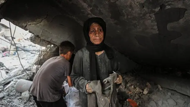 امرأة فلسطينية ترتدي حجاباً داكناً وفستاناً رمادياً، وهي تحمل قطعة ملابس في يدها، تنظر بحزن، بينما تتفقد ما تبقى من الأنقاض في سوق غزة الذي تعرض للقصف الإسرائيلي. ويقف خلفها صبي متجهاً بظهره إلى الكاميرا ويرتدي قميصاً رمادياً قصير الأكمام وبنطالاً أسود.