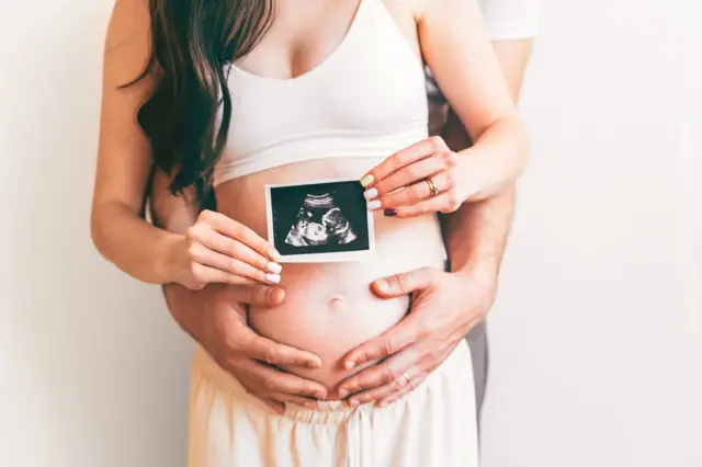 Чоловік обіймає жінку з оголеним животом. В руках вони тримають фото УЗД дитини