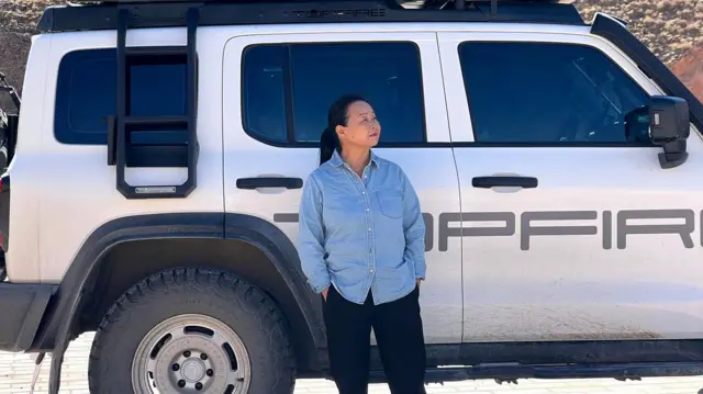 Su Min, vestida con una camisa azul y pantalones negros, parada frente a su hatchback Volkswagen blanco.