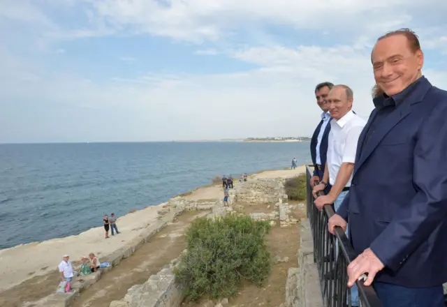 Усміхнені Берлусконі та Путін стоять на оглядовому майданчику над узбережжям біля руїн античного Херсонеса. На передньому плані видно кам’яні залишки стародавніх споруд і море, що простягається до горизонту.