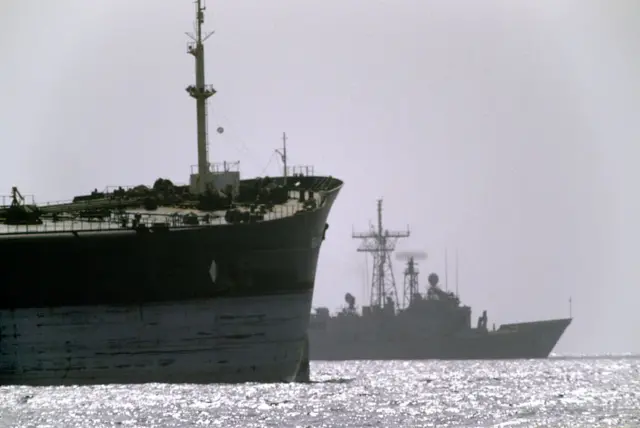 A US warship in the background travels alongside a cargo ship during Operation Earnest Will in the Strait of Hormuz.