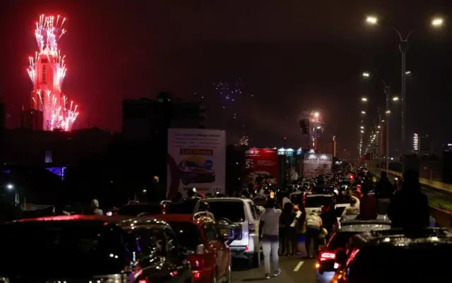 渋滞する道路の向こうで、ピンクにライトアップされた高層ビルから花火が上がっている。道路では車から降りた人たちがそれを見上げている。