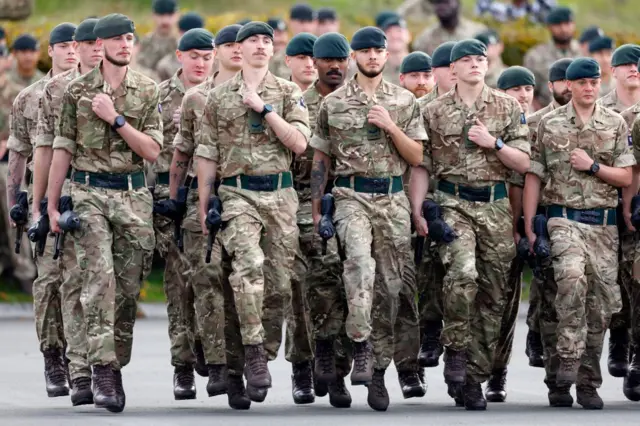 Askerler yan yana geçit töreninde yürüyorlar. Yeşil bereleri var ve yazlık üniformaları giymişler.