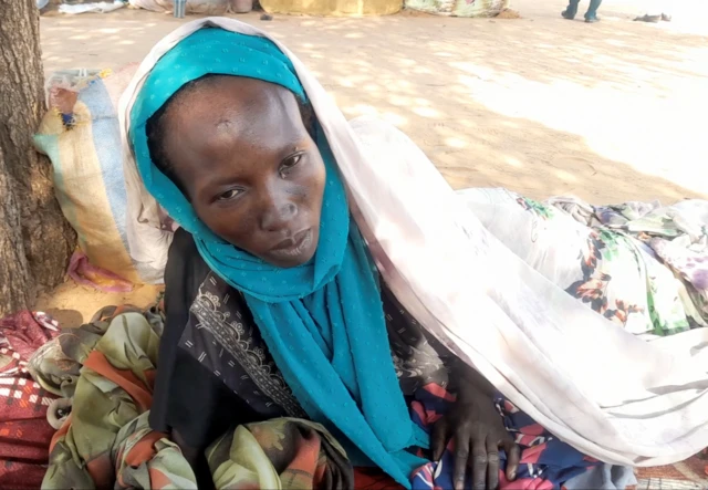 Hamida Adam Ali, vêtue de noir et d'un châle bleu, est allongée sur le sol, recouverte d'une couverture.