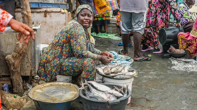 Woman wear colourful printed clothes wit bucket of fish inside market.
