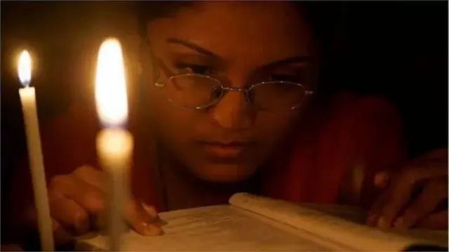 অন্ধকারের মধ্যে দুটি মোমবাতির সামনে বইয়ের দিকে তাকিয়ে আছে চশমা পরা একটি মেয়ে (প্রতীকী ছবি)