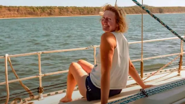 Sarah Packwood está sentadaaposta em aberto betanoum barco,apostaaposta em aberto betanoaberto betanocostas mas olhando para trás para a câmera. Ela está sorrindo, vestida com uma camiseta branca regata e um shorts preto. Em segundo plano, o mar e, mais ao fundo, montanhas.