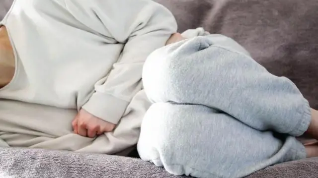A woman in pain laying on a sofa and clutching stomach. Her face is not in the shot.