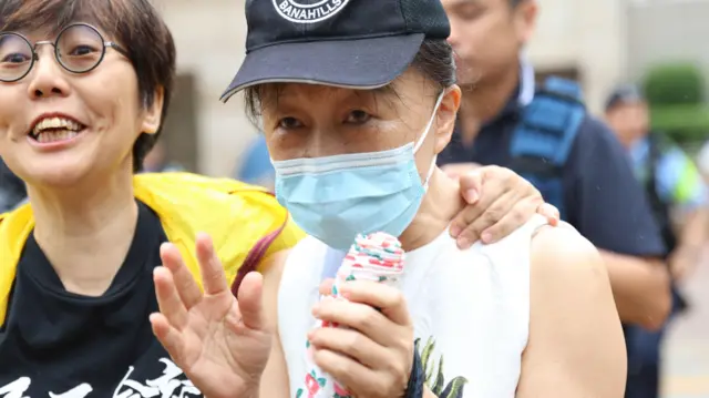 邹幸彤母亲刘华珍(中)在法院外,她戴着蓝色医用口罩和深蓝色鸭舌帽,神情略显慌张(21/8/2026)