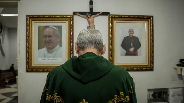 Licio de costas, diante de cruz e imagem do Papa Francisco