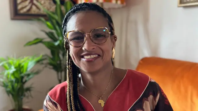 Se ve a Sona Barbosa con gafas y sonriendo mientras está sentada en el interior, con plantas de interior y un sofá visibles detrás de ella.