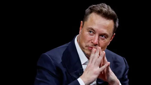 白いシャツと紺色ブレザーを着て座っているイーロン・マスク氏が、両手の指を口の前で合わせて、右斜め前を見ている。