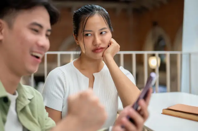 An unhappy, moody young Asian woman is feeling jealous, secretly casting a suspicious look at her boyfriend as he texts on his smartphone while they hang out at a coffee shop.