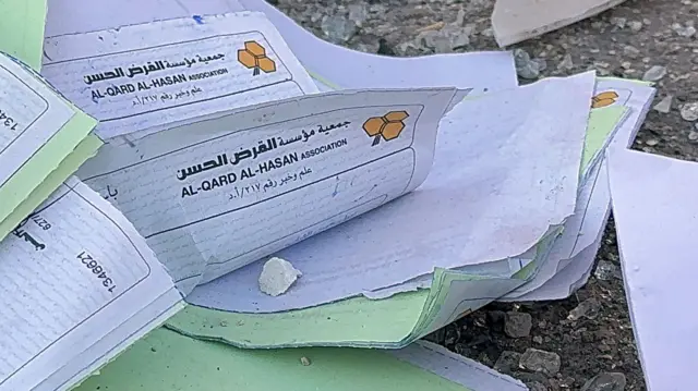 اسناد پخش شده روی زمین که روی آنها «القرض‌الحسن» نوشته شده است، در خارج از یکی از شعبه‌های آسیب دیده پس از حملات هوایی اسرائیل۲۰ اکتبر ۲۰۲۱ که چندین شعبه از این موسسه مالی را هدف قرار داد