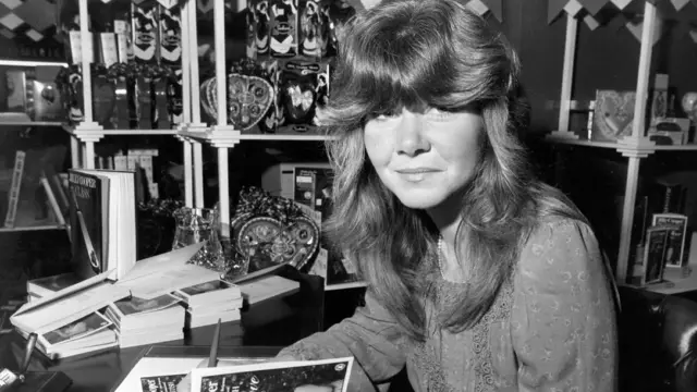 Чорно-біле фото Джиллі Купер, вона сидить за столом з ручкою і дає автографи