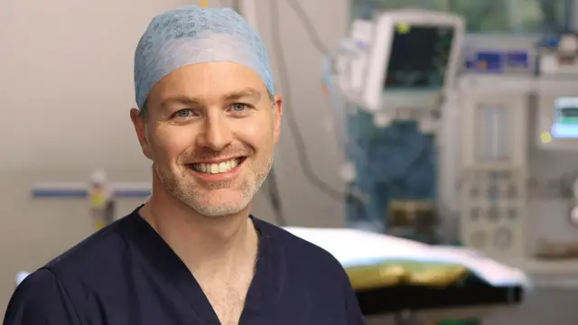 一名男子在手術室中微笑，頭戴髮網並穿著深藍色醫療手術服