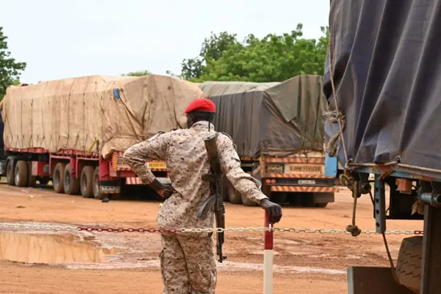 Un homme en treillis avec une arme à côté des véhicules longs chargés de marchandises