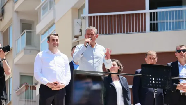 Özgür Özel otobüs üzerinde konuşuyor. Yanında eşi ve takım elbiseli kişiler var.