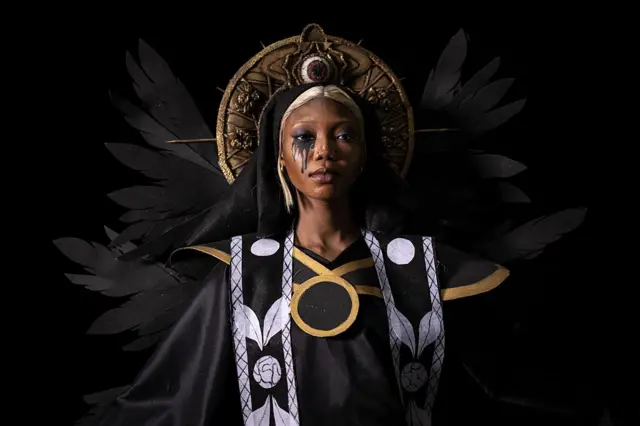A woman poses for the camera wearing an imposing black and white dress and golden headdress.