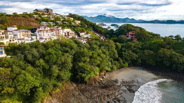 Una colina con árboles frondosos, casas blancas con tejas y una playa en frente. En la distancia se ve más mar y montañas
