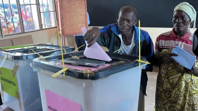 Umugabo n'umugore bariko bashira impapuro batoreyeko mu masandugu y'itora. Iyiriko igipapuro c'urwatsi rubisi ni iyo amatora y'abashingamateka, nayo iryo ibara ry'iroza ni iyo abajenama ba komine. Inyuma yabo, haboneka uwundi muntu yambaye ishati yera, akiriko aratora mu bwiherero (uyu ni perezida Evariste Ndayishimiye) 