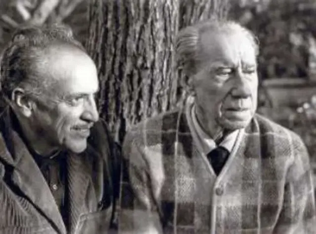 Dos hombres mayores, vestidos casualmente, sentados frente a un árbol