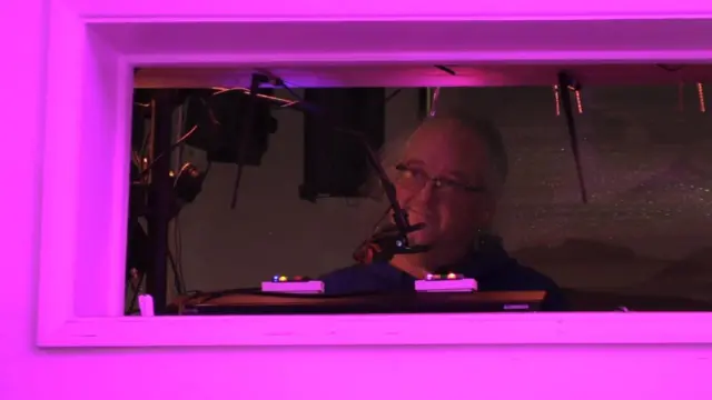Mirando a Charles en su cabina de sonido, enmarcado por la ventana y la iluminación rosa.