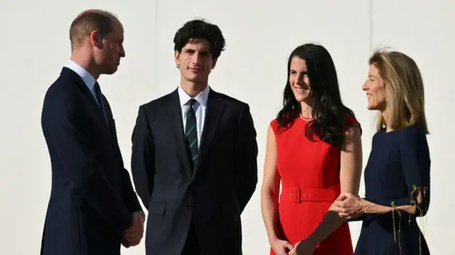 Prince William dey stand and chat wit Jack, Caroline and Tatiana Schlossberg. All of dem wear very dark suits and Tatiana wear bright red dress.