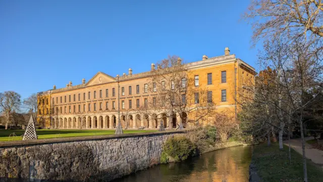 Un gran edificio de piedra amarilla de tres plantas junto a un pequeño río. Hay un césped bien cuidado frente al edificio. El cielo es azul, sin nubes, y el edificio está bañado por la luz del sol.