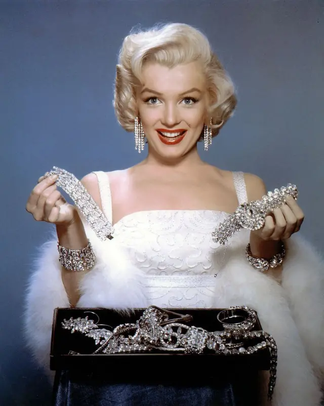Una sonriente Marilyn Monroe sostiene joyas en ambas manos y tiene un joyero delante