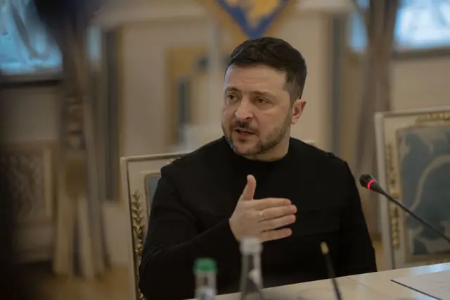 Зеленський у чорній кофті сидить за столом в урочистому приміщенні. Перед ним – мікрофон і кілька пляшок з водою, а рука жестикулює під час розмови.
